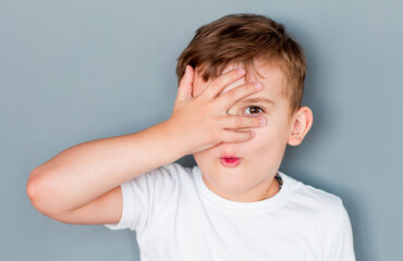 Young boy closed one of his eye with surprised expression on a gray background, shocked amazed terrified gesture