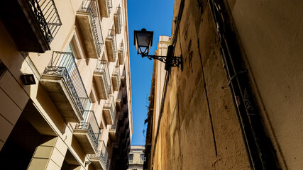 Fototapeta premium Low-angle view of European narrow alley with balconies and a vintage street lamp under a clear blue sky, evoking Mediterranean architecture