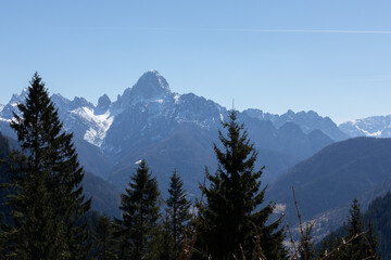 vista dettagliata di una catena montuosa lontana, con le sue cime e i pendii parzialmente innevati, vista da dietro gli alberi di un bosco di montagna, di giorno, in primavera, con cielo sereno