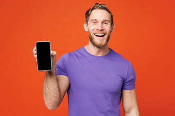 Young smiling happy man he wear purple t-shirt casual clothes hold in hand use mobile cell phone with blank screen workspace area isolated on red orange background studio portrait. Lifestyle concept.