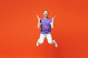 Full body young surprised shocked excited happy man he wearing purple t-shirt casual clothes jump high spread hands looking camera isolated on red orange background studio portrait. Lifestyle concept.