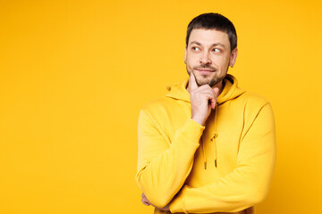 Young minded pensive Caucasian man wears hoody casual clothes put hand prop up on chin, lost in thought and conjectures isolated on plain yellow orange background studio portrait. Lifestyle concept