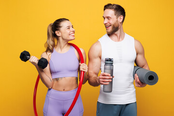 Young strong fitness trainer instructor sporty two man woman wearing blue clothes spend time in home gym hold yoga mat dumbbells hula hup isolated on plain yellow background Workout sport fit concept
