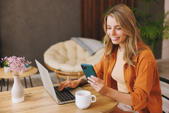 Sideways young smart IT woman wear orange shirt use laptop pc computer hold mobile cell phone sit alone at table in coffee shop cafe restaurant indoors work or study Freelance office business concept.