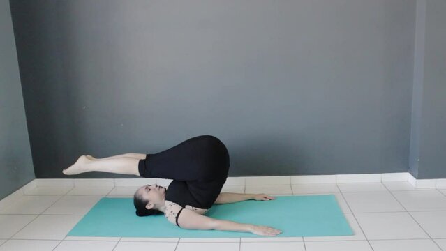 Female fitness instructor performing halasana sarvangasana baddha konasa pose variations on yoga mat. Woman training yoga in workout practice studio.