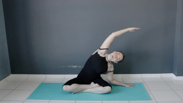 Female fitness instructor performing a body side stretching movement, sit on semi lotus pose. Siddhasana yoga posture.