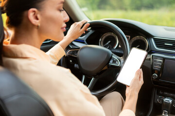 No cell phone use while driving car. Young lady holding cellphone and steering wheel, sitting in own car, phone with blank white screen for ad