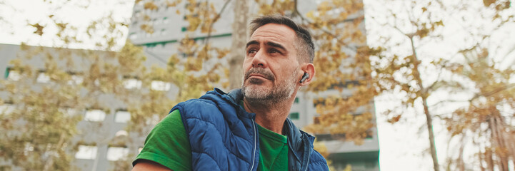 Mature man wearing casual clothes in wireless earphone enjoying music while sitting on park bench, bottom view, soft focus