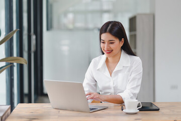 Asian businesswoman in white blouse pondering over new project, working on laptop in a contemporary office space.