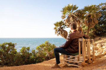 Couple sitting on a wooden bench overlooking the ocean at sunset. They are embracing and enjoying a moment of tranquility and connection in a scenic natural setting with palm trees.