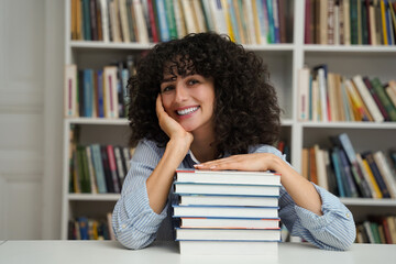 Cheerful student girl sitting at desk with stack of books in library