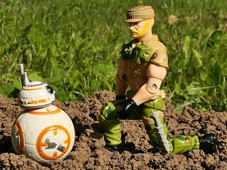 Fototapeta premium GI JOE action figure of infantry soldier Rocknroll from year 1989 meets model of Star Wars spherical astromech droid BB8 on cultivated spring garden soil, green lawn in background. 