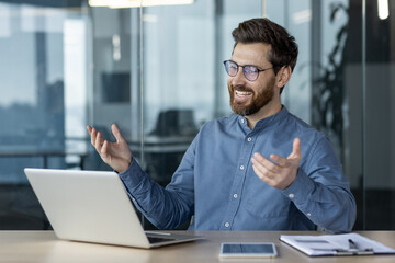 Smiling young man businessman sitting in the office at the desk and talking on a video call on the...