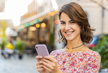 Smiling happy young woman using smartphone typing text messages browsing internet, finishing work, looking at camera outdoors. Adult girl tourist standing in urban city street outside. Town lifestyles