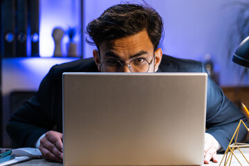 Confident Indian businessman hiding behind laptop computer, looking at camera, spying his colleagues working, peeping. Professional freelancer man looking from behind computer, cunning eyes glance