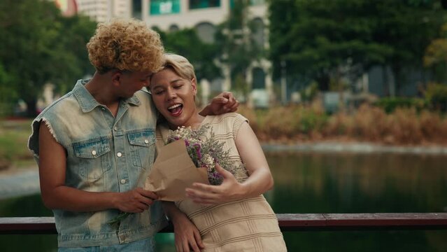 Romantic gay boyfriend presents flower bouquet to happy man on urban park bridge. Homosexual couple celebrates anniversary in garden. LGBT community people visibility in pride month