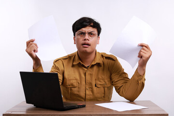 Stressed male teacher in civil servant uniform with paper and laptop on the desk holding papers