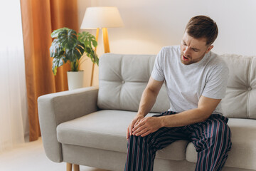 Unhappy man sitting on sofa at home and touching hurt knee with grimace of pain and suffering. Young guy with rheumatism, arthritis or osteoarthritis feeling intense severe pain in knee joint