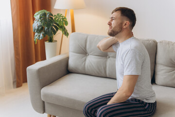 Fototapeta premium Neck Pain. Caucasian Man Massaging Aching Neck On Couch In Living Room At Home. Osteoarthritis, Health Problem Concept.