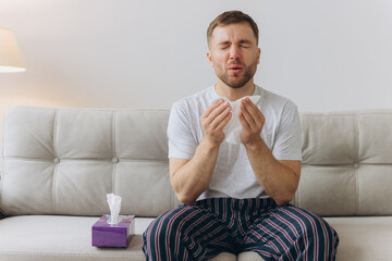 Unhappy middle aged man suffering from fever and flu, blowing nose in napkin, sitting in living room interior. Covid-19 lockdown, treatment of illness, cold and runny