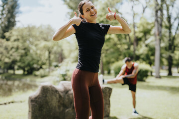 Obraz premium Active young woman in athleisure gives a thumbs up while working out in a sunny park, with a male friend stretching in the background.
