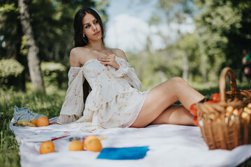 A young, stylish woman relaxes on a picnic blanket in the park, surrounded by a basket of fresh...