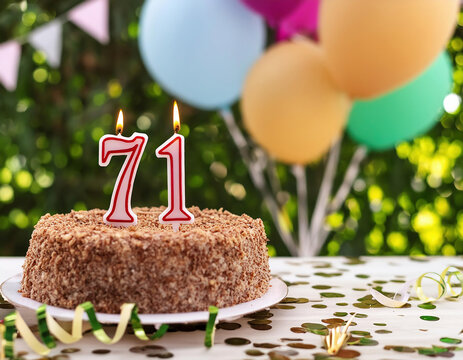 A birthday celebration cake adorned with burning candles with number 71 age, balloons and party ornaments against a green garden	