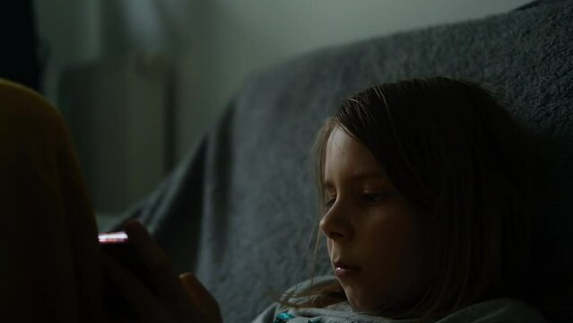 A young girl scrolls through her phone in the dim light of her bedroom, capturing a relatable moment of late-night screen time
