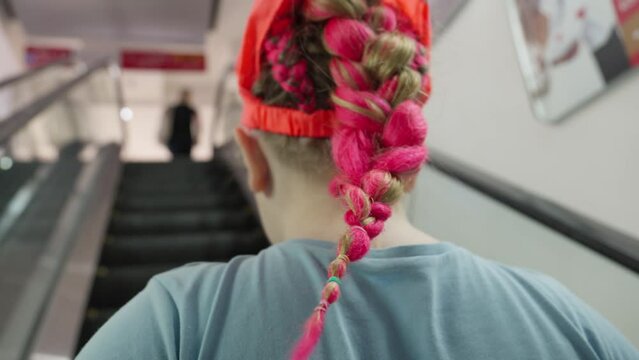 Girl with pink braided hair riding escalator. Back view. She enjoying her day out.