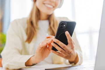 Young professional woman listening music using headphones and browsing smartphone at home office