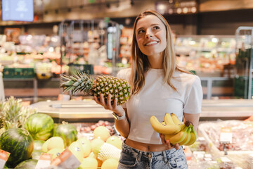 Young woman choosing between pineapple and banana while shopping for groceries in supermarket