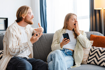 Young woman yawning while holding smartphone and sitting next to man talking to her
