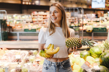 Young woman holding pineapple and bananas, smiling at the grocery store
