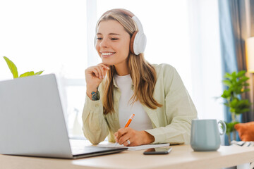 Young woman wearing headphones taking notes while attending online class