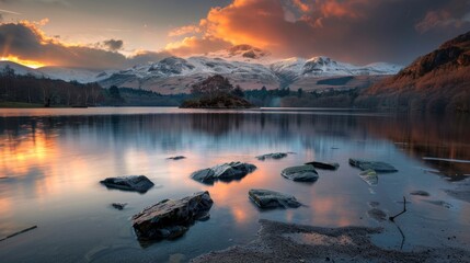 A serene lake reflects the vibrant colors of a stunning sunset, with snow-capped mountains and a small island in the background. Rocks are scattered in the foreground. - Powered by Adobe