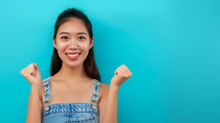 Asian woman with long dark hair wearing blue jean overall with happy expression on face raising fists in victory joy,