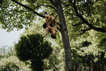 Young man performing acrobatic stunt by hanging upside down from a tree branch in a lush green forest.