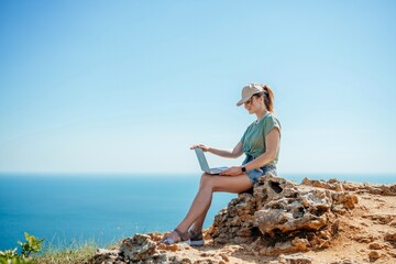 A woman is sitting on a rock overlooking the ocean with a laptop in front of her