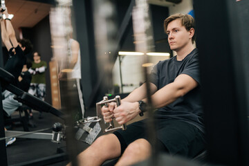 Side view of focused motivated sportsman working out on stationary rowing machine in gym. Young...