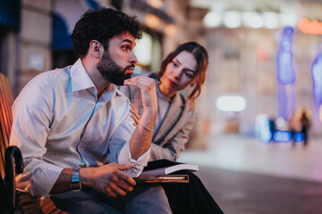 Obraz premium Two businesspeople having a thoughtful conversation on a bench in an urban area at night, deeply engaged in discussion.