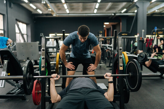 Front view of experienced fitness instructor helping beginner sportsman doing barbell bench press exercise during personal workout in gym. Male doing barbell bench press under couch supervision