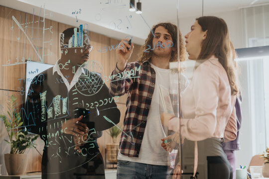 A multi-ethnic group of business workers are engaged in a brainstorming session, using sticky notes and markers to write on a glass wall, analyzing data and strategizing.