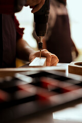 Craftsperson drills hole into lumber block, doing furniture assembling in carpentry shop, close up shot. Manufacturer in assembly shop inserting nail into wood plank using electric power drill