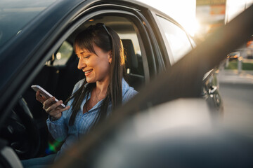 Trendy woman sitting in her opened car and smiling at her phone.