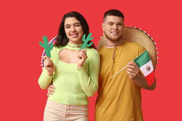 Beautiful young happy couple with sombrero hats, paper cacti and Mexican flag on red background. Cinco de Mayo celebration