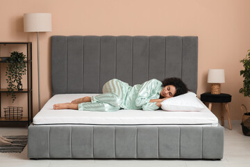 Young African-American woman with pillow lying on soft mattress in bedroom