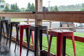 Four colorful iron metal stackable bar stools; black, and red colored with footrests. The chairs are on a wooden restaurant patio. There's a lush green fenced-in grassy field off the porch.  