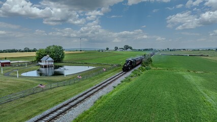 A Vintage Steam Train Travels Along A Railroad Track Through Lush Green Farmland, Passing By A Small Pond With A Charming House, Under A Sky Filled With Puffy Clouds In A Serene Rural Landscape.