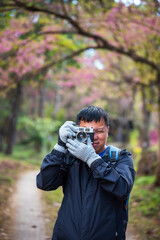 Vertical image Asian man use professional digital camera take a photo. Smiling male photographer look at photo film camera outdoors. Man shooting photo in green nature park. Men hobby lifestyle