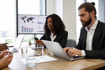 Diverse business team engaged in serious discussion using electronic devices during meeting room presentation, digital charts on screen.